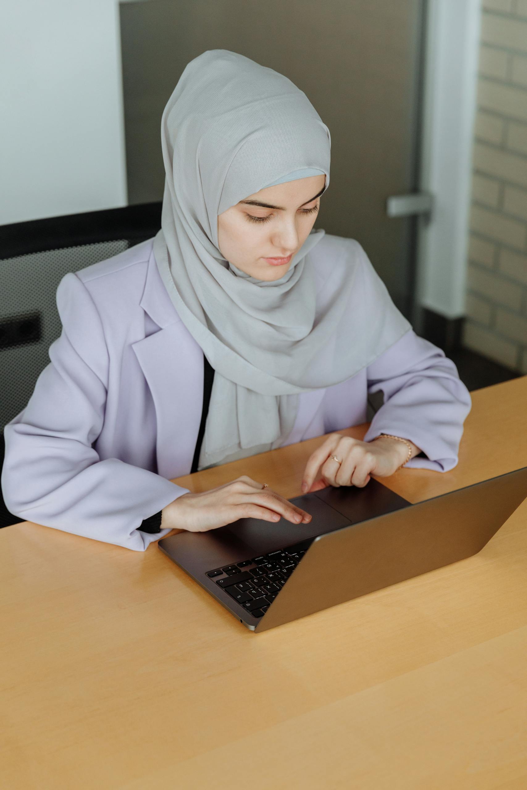 Muslim woman in hijab typing on a laptop in a modern office setting.