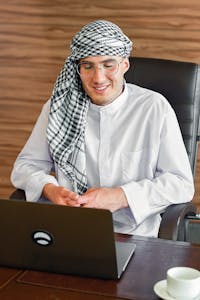 Smiling Middle Eastern man in traditional attire working on a laptop indoors.